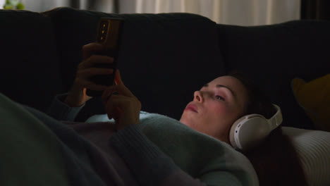 Woman-Wearing-Wireless-Headphones-Lying-On-Sofa-At-Home-At-Night-Streaming-Music-Or-Watching-Movie-On-Mobile-Phone-1